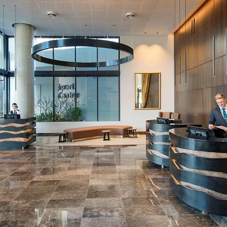 Hotel Pullman Brisbane Airport Exterior foto The image shows a modern hotel lobby. There are two reception desks made of dark-colored materials, and staff members are attending to guests at these desks. The lobby features large windows that allow natural light to fill the space, and there are p