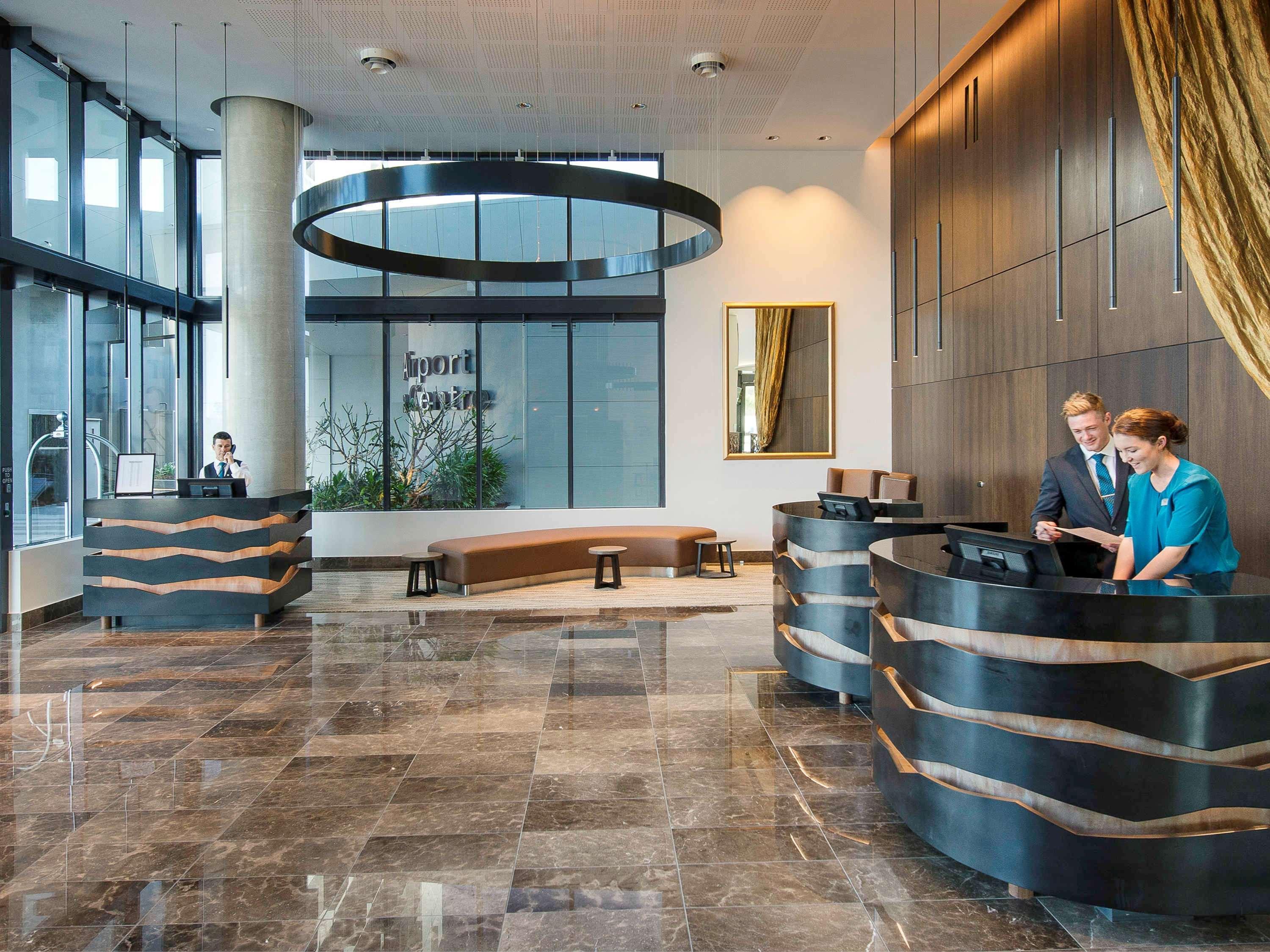 Hotel Pullman Brisbane Airport Exterior foto The image shows a modern hotel lobby. There are two reception desks made of dark-colored materials, and staff members are attending to guests at these desks. The lobby features large windows that allow natural light to fill the space, and there are p